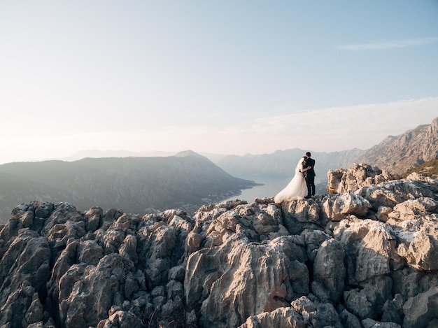 La novia y el novio se abrazan en una montaña rocosa y miran la vista posterior de la bahía