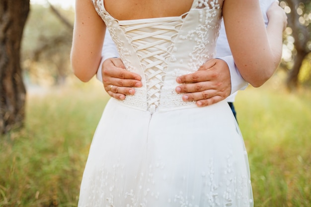 La novia y el novio se abrazan en un olivar, las manos del novio en el primer plano de la cintura de la novia
