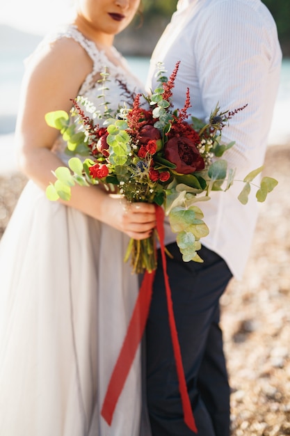 La novia y el novio se abrazan y sostienen el ramo de la boda en la playa de guijarros junto al mar