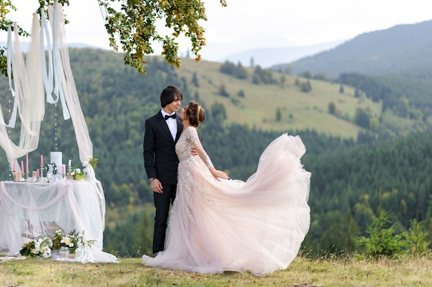 La novia y el novio se abrazan bajo un viejo roble. Sesión de fotos de boda en las montañas. Junto a ellos se prepara una decoración para la ceremonia.