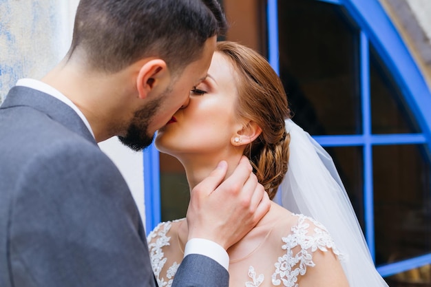 La novia y el novio amorosos jóvenes se abrazan y besan cerca de las ventanas azules