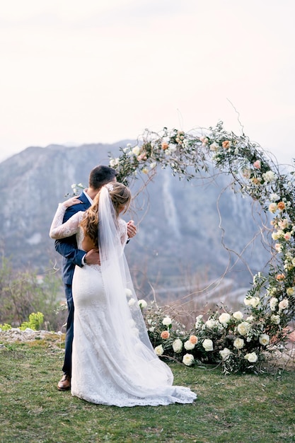 La novia y el novio bailan abrazándose y miran a través del arco de la boda hacia las montañas.