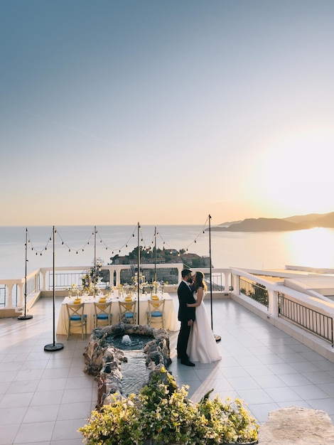 La novia y el novio se besan en la terraza del restaurante cerca de la mesa puesta y la fuente