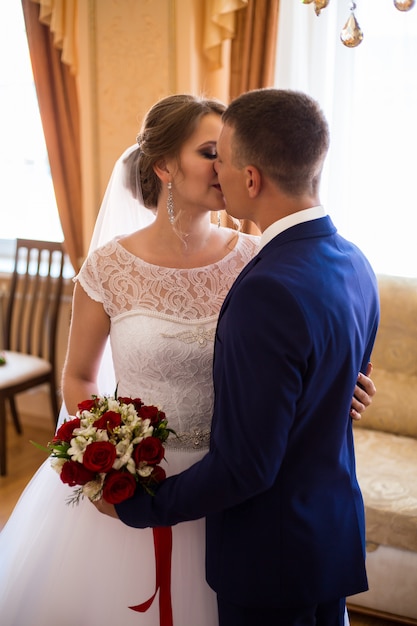 La novia y el novio besándose en la habitación del hotel, sosteniendo un ramo de novia