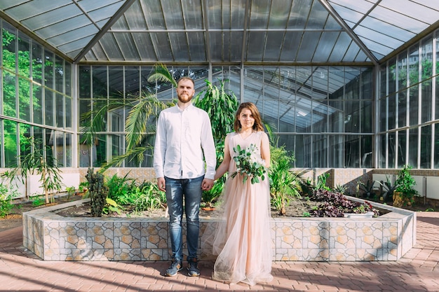 La novia y el novio caminan en el jardín botánico cerca de la antigua arquitectura de vidrio tomándose de la mano