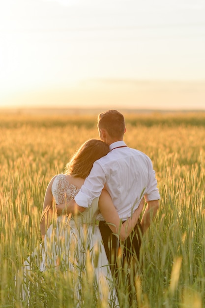 Novia y el novio en un campo de trigo. La pareja se abraza durante el atardecer y mira a la distancia de espaldas a la cámara.