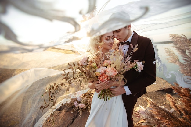 La novia y el novio cerca de la decoración de la boda en una ceremonia en un acantilado de roca cerca del agua al atardecer. Velo volando del viento