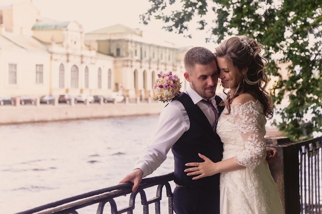 La novia y el novio están abrazando la orilla del río con vistas a los edificios históricos