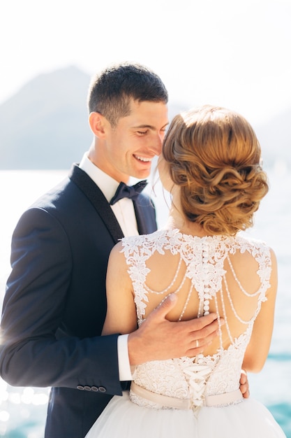 La novia y el novio están de pie abrazándose y sonriendo en el muelle cerca del primer mar