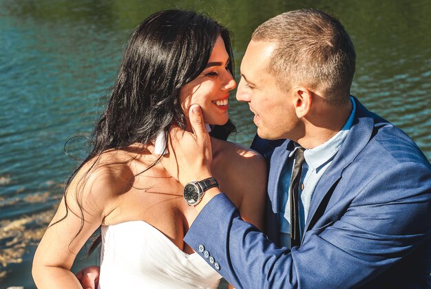 La novia y el novio están de pie junto al río, uno frente al otro y besándose en el parque en un día soleado