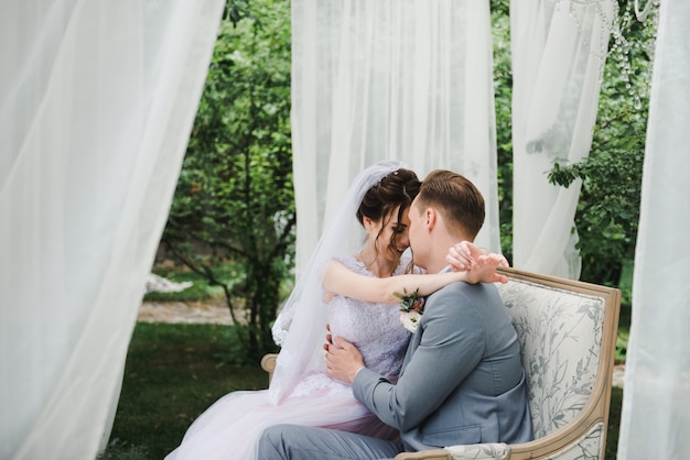 La novia y el novio están sentados en un hermoso sofá en un mirador en el jardín