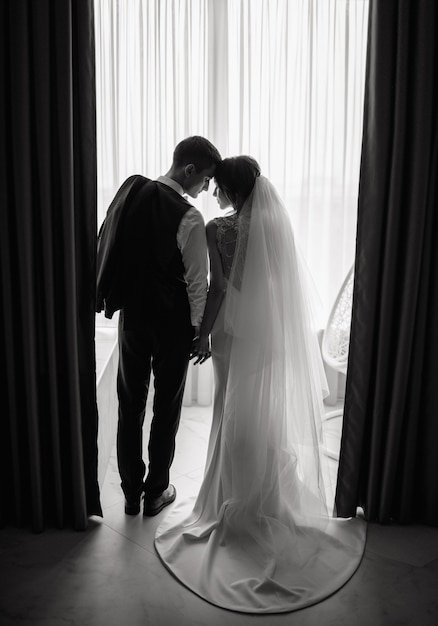 La novia y el novio están tomados de la mano frente a la ventana con las cabezas inclinadas en el hotel. Boda, amor, concepto de relación. Clave baja. Retrato de cuerpo entero.