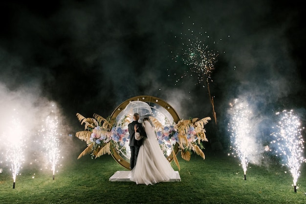 La novia y el novio jóvenes se paran cerca del arco de la boda por la noche con luces de humo y siluetas de fuegos artificiales de los recién casados