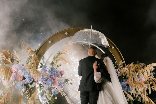 La novia y el novio jóvenes se paran cerca del arco de la boda por la noche con luces de humo y siluetas de fuegos artificiales de los recién casados