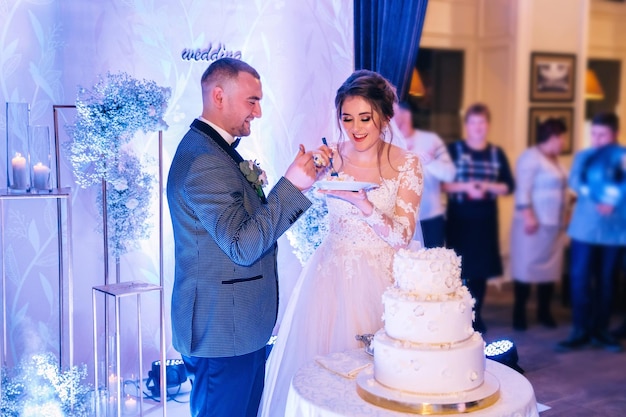 La novia y el novio juntos cortan un pastel blanco de bodas y se alimentan mutuamente en un ambiente romántico