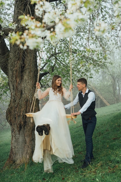 La novia y el novio montan un columpio de cuerda en el jardín. Boda en el bosque.