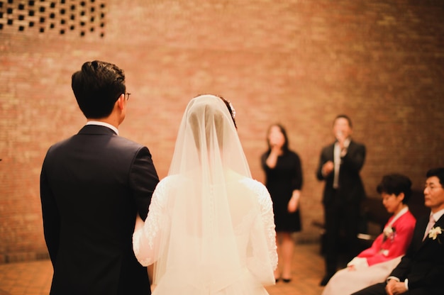 Una novia y un novio se paran frente a una pared de ladrillos y el novio lleva un vestido de novia blanco.