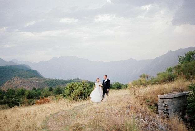 La novia y el novio tomados de la mano caminan por un camino de tierra en las montañas