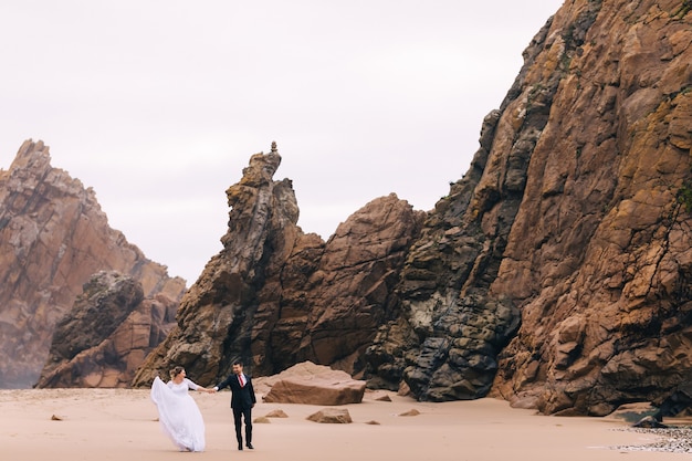 La novia y el novio tomados de la mano y caminar sobre la arena con un fondo de acantilados de piedra se divierten