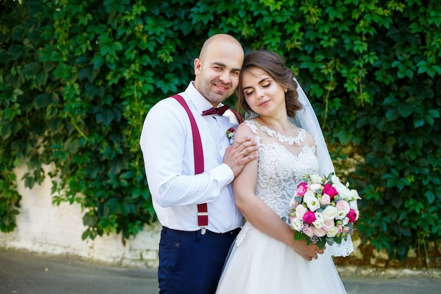 Novia pareja alegre con un vestido blanco con un ramo mientras el novio con tirantes y pajarita. En el contexto de una pared con hojas verdes. Pareja feliz. El concepto de matrimonio.