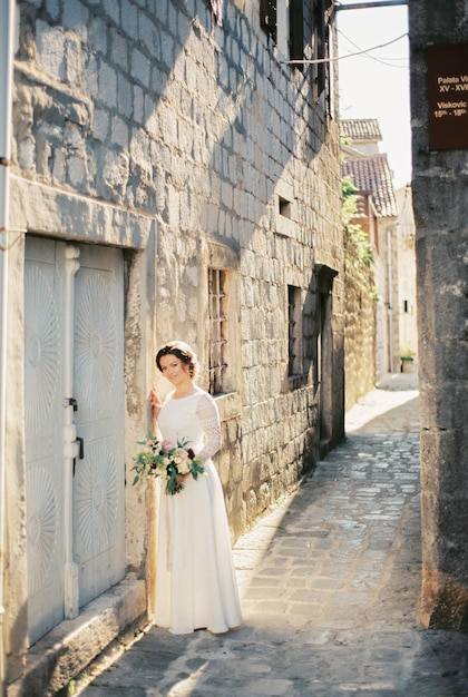 La novia con un ramo se encuentra cerca de la pared de piedra de una casa antigua en una calle estrecha