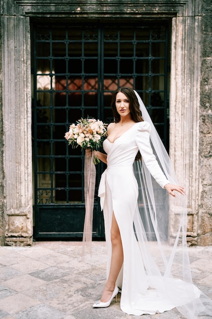 Foto la novia con un ramo de flores está de pie cerca de la puerta de celosía de un edificio antiguo sosteniendo su velo