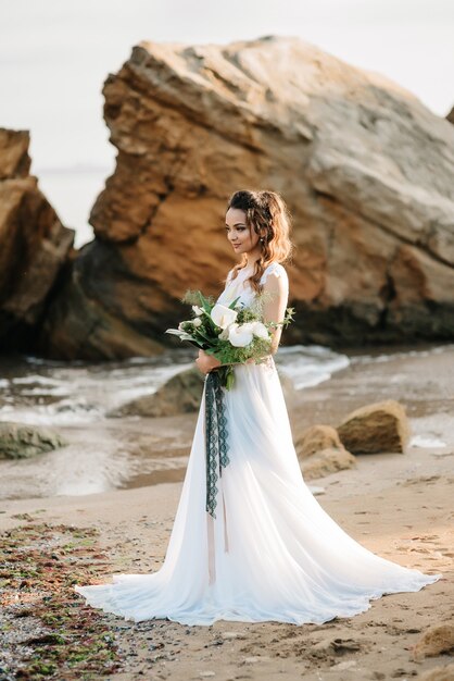 Novia con un ramo de novia en la orilla del mar negro a la luz del atardecer