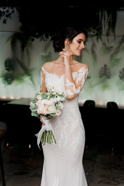 Novia con ramo de novia sonríe y toca su rostro y cabello. Chica en vestido de novia en restaurante de moda.