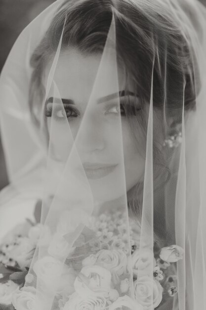Novia rubia rizada en un vestido blanco cubierto con un velo posa para la cámara con un ramo de rosas Retrato de la novia hermoso maquillaje y cabello Wedding in nature foto en blanco y negro