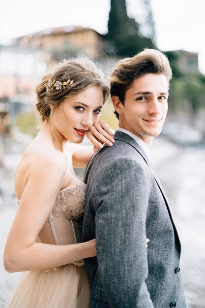 La novia sonriente abraza al novio desde atrás mientras está de pie en el paseo marítimo del lago como