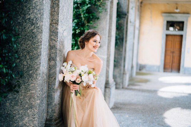 Novia sonriente con un vestido con un ramo de flores rosas se sentó cerca de un pilar en una habitación abovedada