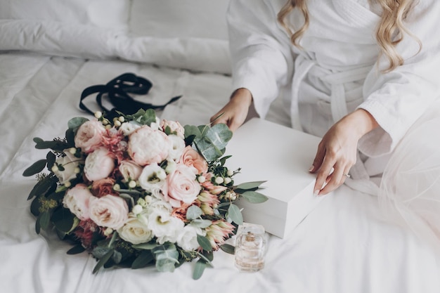 Novia sosteniendo una caja blanca para el tocador de la mañana antes de la ceremonia de la boda Ramo de boda moderno botella de perfume y caja de regalo en la cama blanca Arreglos de boda o despedida de soltera