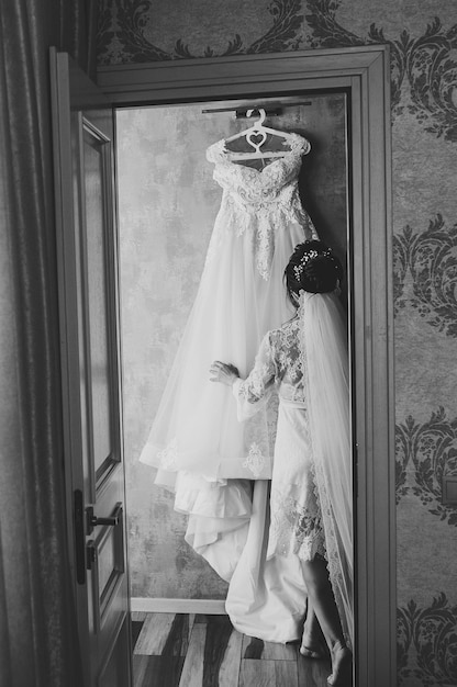 Novia sosteniendo un vestido en la habitación chica con un vestido de noche sexy en casa Preparándose para una boda en la mañana Vista posterior Foto en blanco y negro