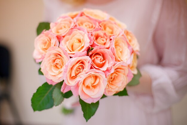 La novia sostiene un ramo de flores rosadas con hojas verdes en sus manos. Primer plano, interior