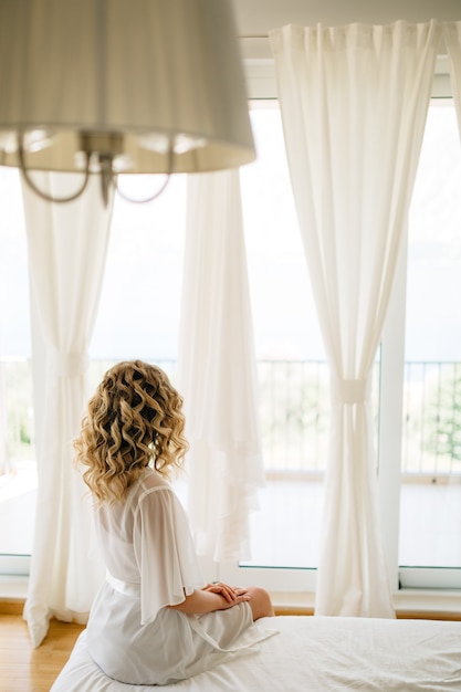 Foto una novia en una suave bata sentada en la cama junto a la amplia ventana de la terraza con cortinas blancas