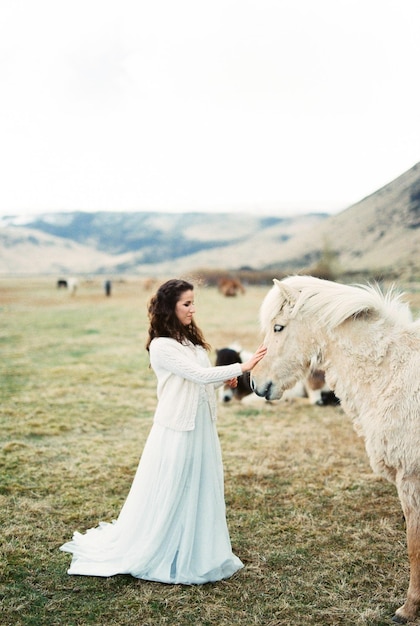 La novia con un vestido blanco acaricia un caballo beige en el césped Islandia