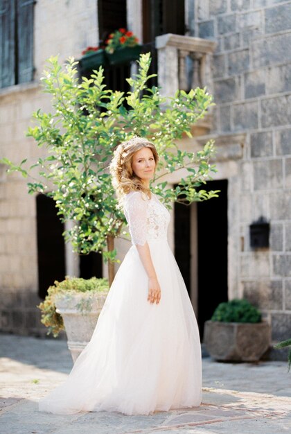 La novia con un vestido blanco se encuentra cerca de un árbol en una tina en el patio