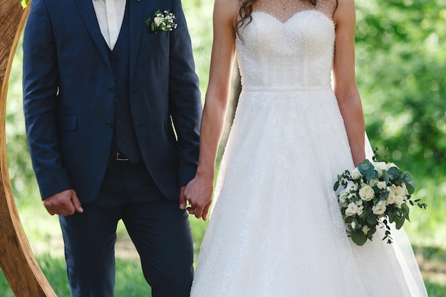 Foto novia en vestido blanco con hermoso ramo y novio tomados de la mano al aire libre en la ceremonia de la boda