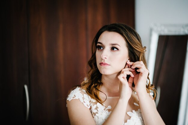 La novia con un vestido blanco lleva pendientes de oro con perlas. En las manos de la novia decoración en forma de pendiente. Mañana de bodas. Joyería.