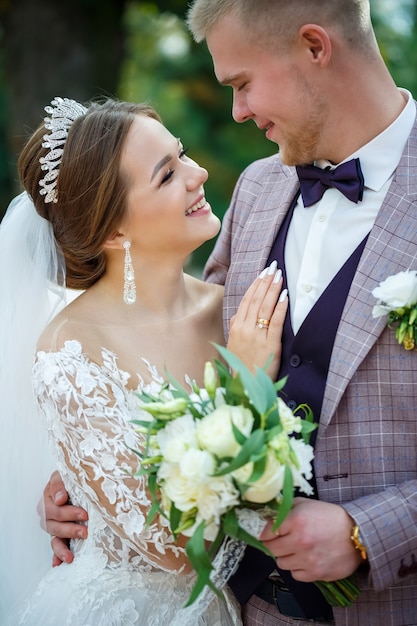 La novia en vestido blanco y el novio en traje se abrazan y caminan en el parque