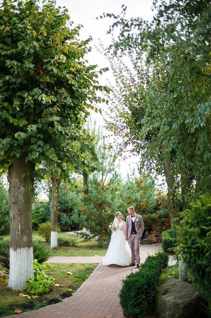 La novia en vestido blanco y el novio en traje se abrazan y caminan en el parque