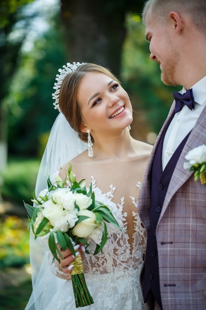 La novia en vestido blanco y el novio en traje se abrazan y caminan en el parque