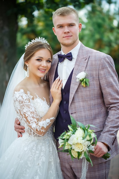 La novia en vestido blanco y el novio en traje se abrazan y caminan en el parque