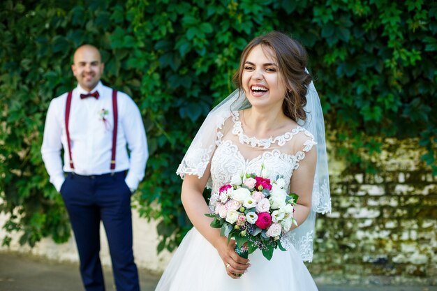 La novia con un vestido blanco en primer plano y el novio con tirantes detrás. en la pared de fondo con hojas verdes