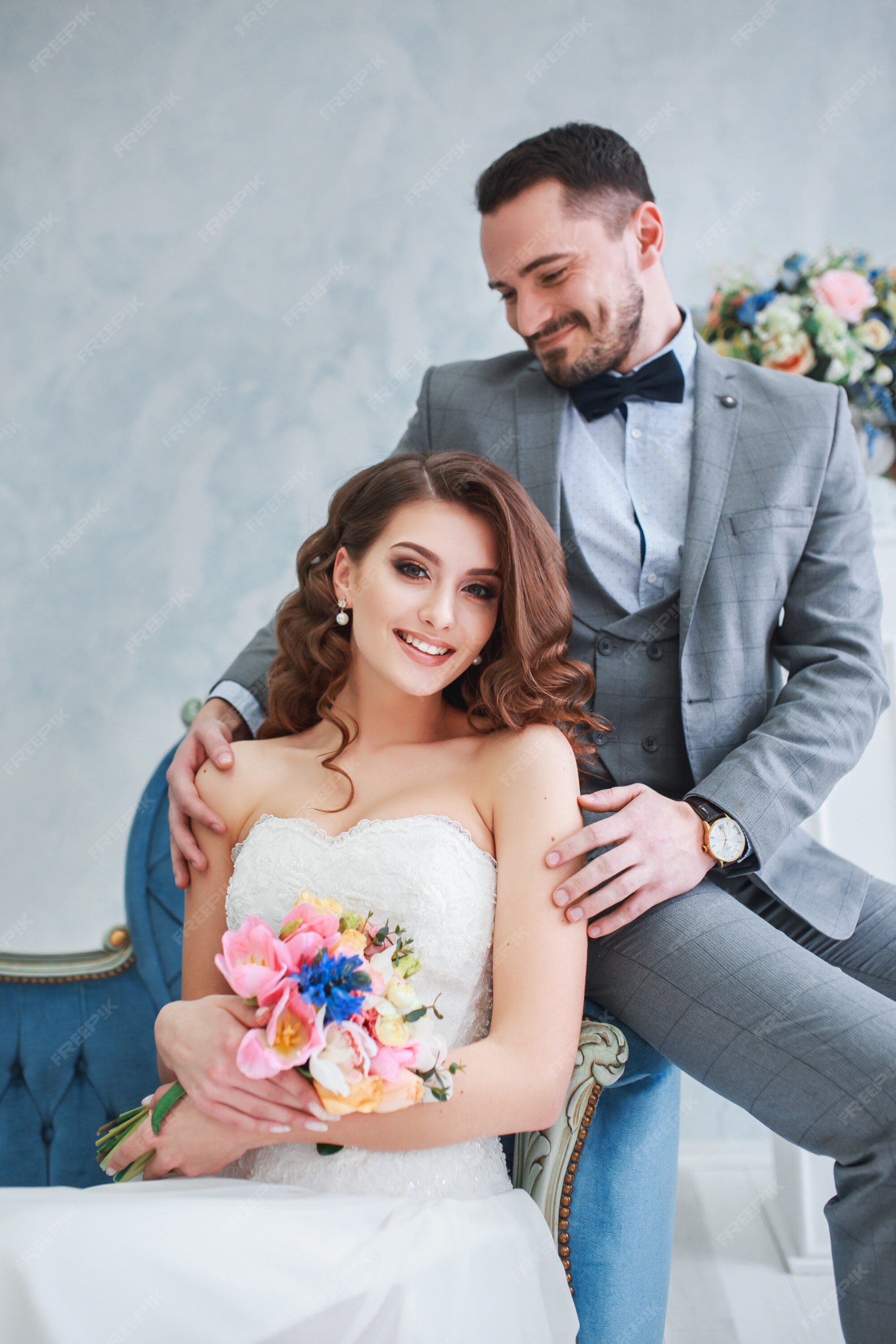 Novia en vestido hermoso y en traje gris sentado en el sofá en el interior. estilo de boda de moda | Foto Premium