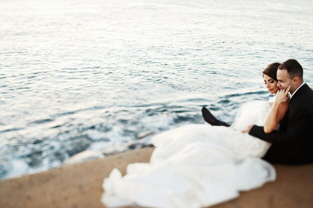 Foto la novia con un vestido magnífico yace sobre las rodillas del novio mientras descansan en la orilla del océano