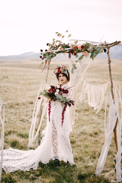 Novia con un vestido de mimbre y una corona con un ramo de flores se encuentra cerca del arco de la boda