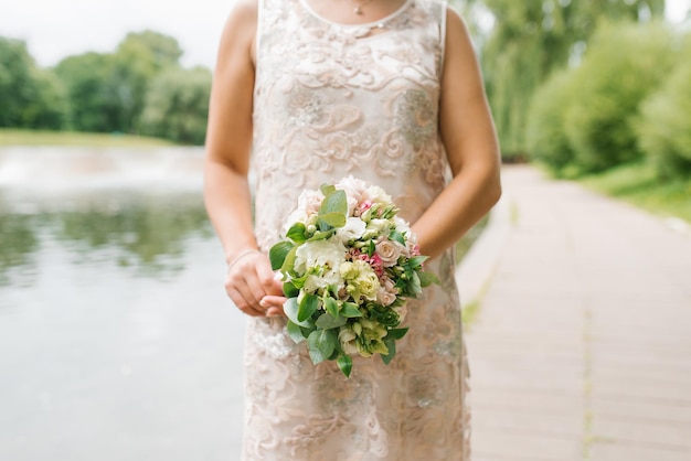 Una novia con un vestido de novia beige sostiene un ramo de flores y vegetación en sus manos.