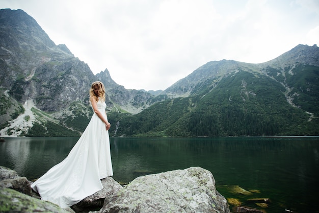 La novia con un vestido de novia blanco se para y mira las montañas y el lago.