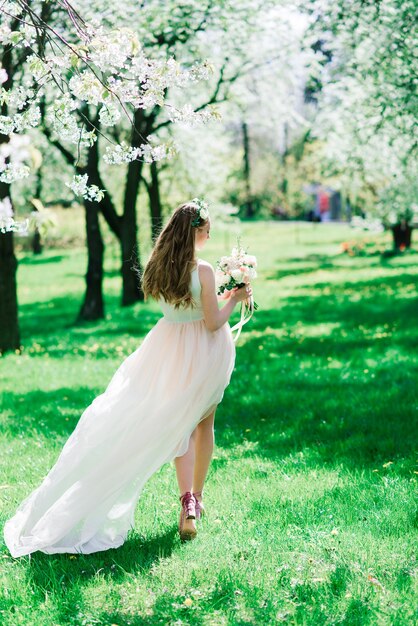 La novia en vestido de novia blanco sostiene un ramo en el parque verde